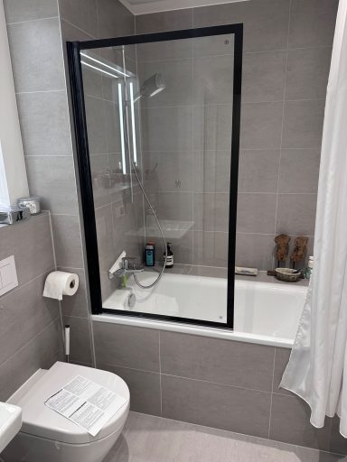 Modernes Badezimmer mit Badewanne, Dusche und einem WC, grauen Fliesen und weißer Duschvorhang.